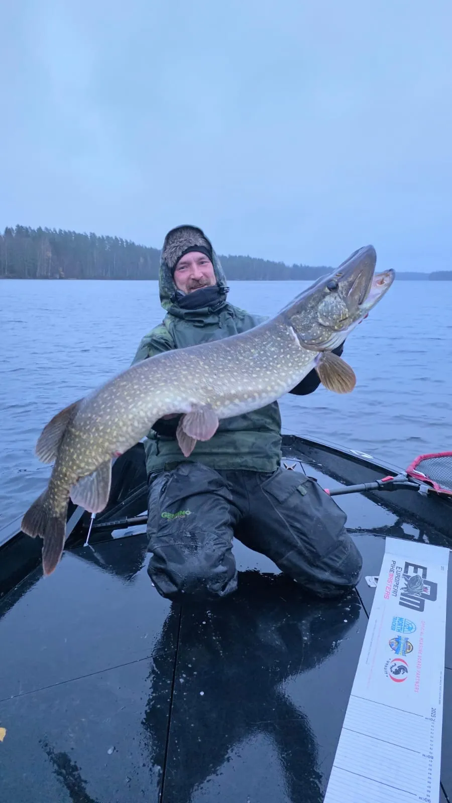 Mies polvistuneena veneessä pitää suurta haukea ulkona järvellä.
